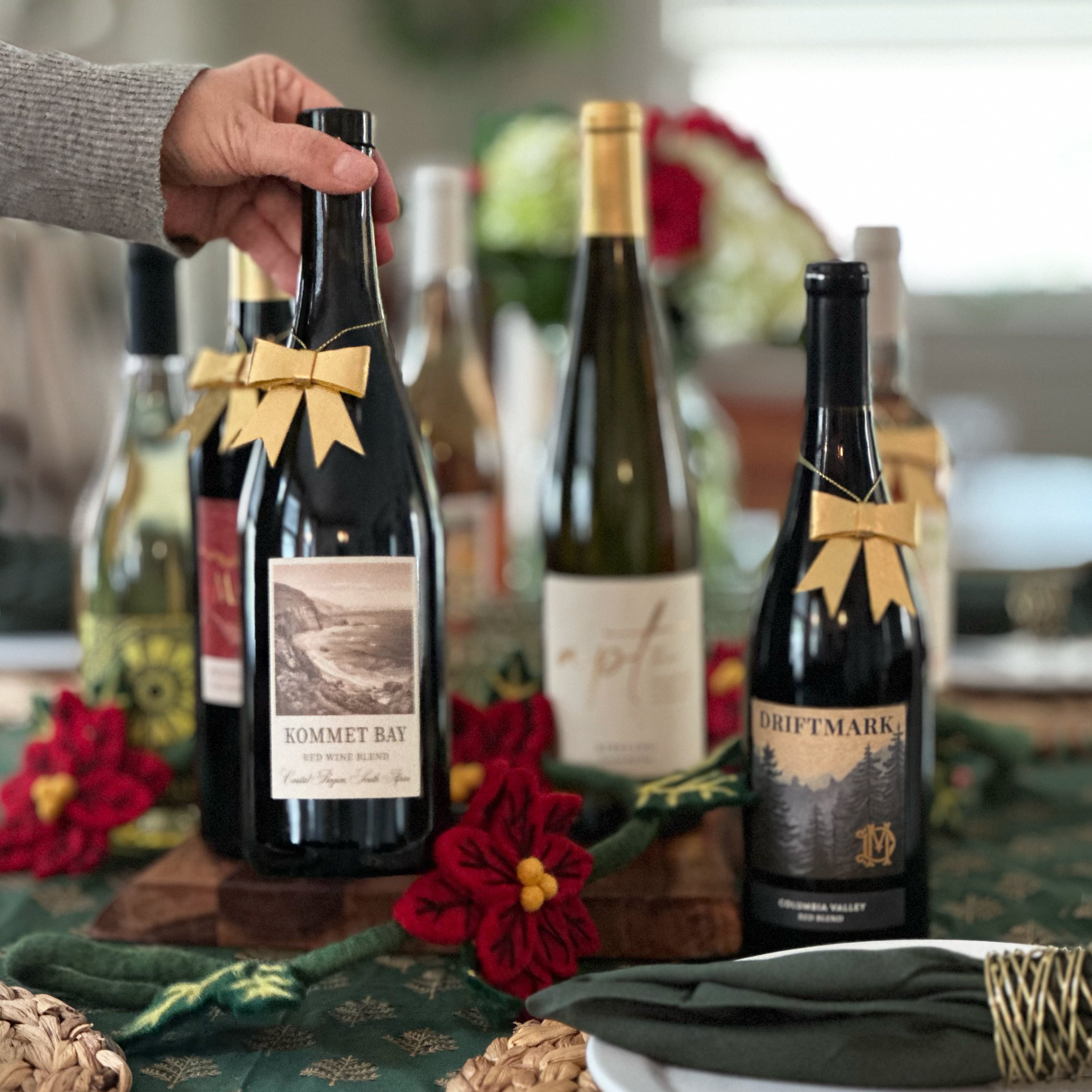 Hand placing a wine bottle with a gold bow among a selection of wine bottles on a festive table with holiday decorations