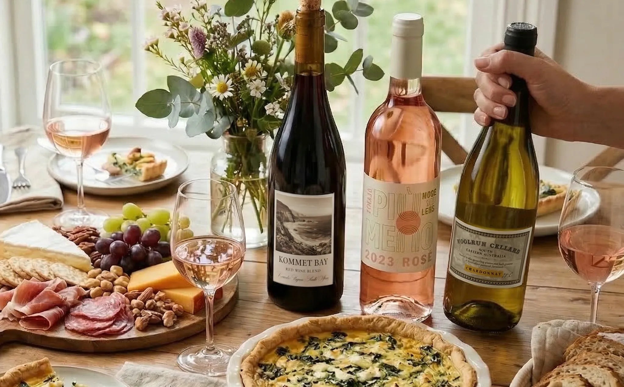 Red, white, rose wines on a wooden table with cheese, grapes, salami, and quiche, a hand reaching for the white wine