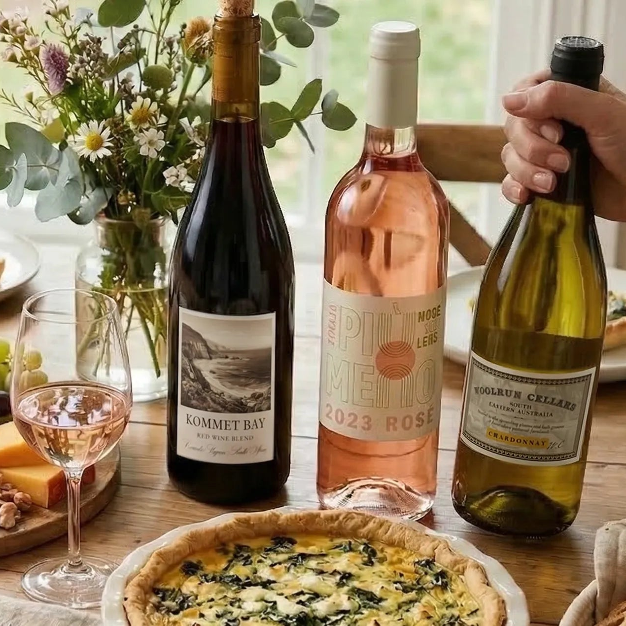 Easter wine (red, white, rose) on a table with a spinach quiche, cheese and a flower vase in the background. A hand reaching for the white wine.