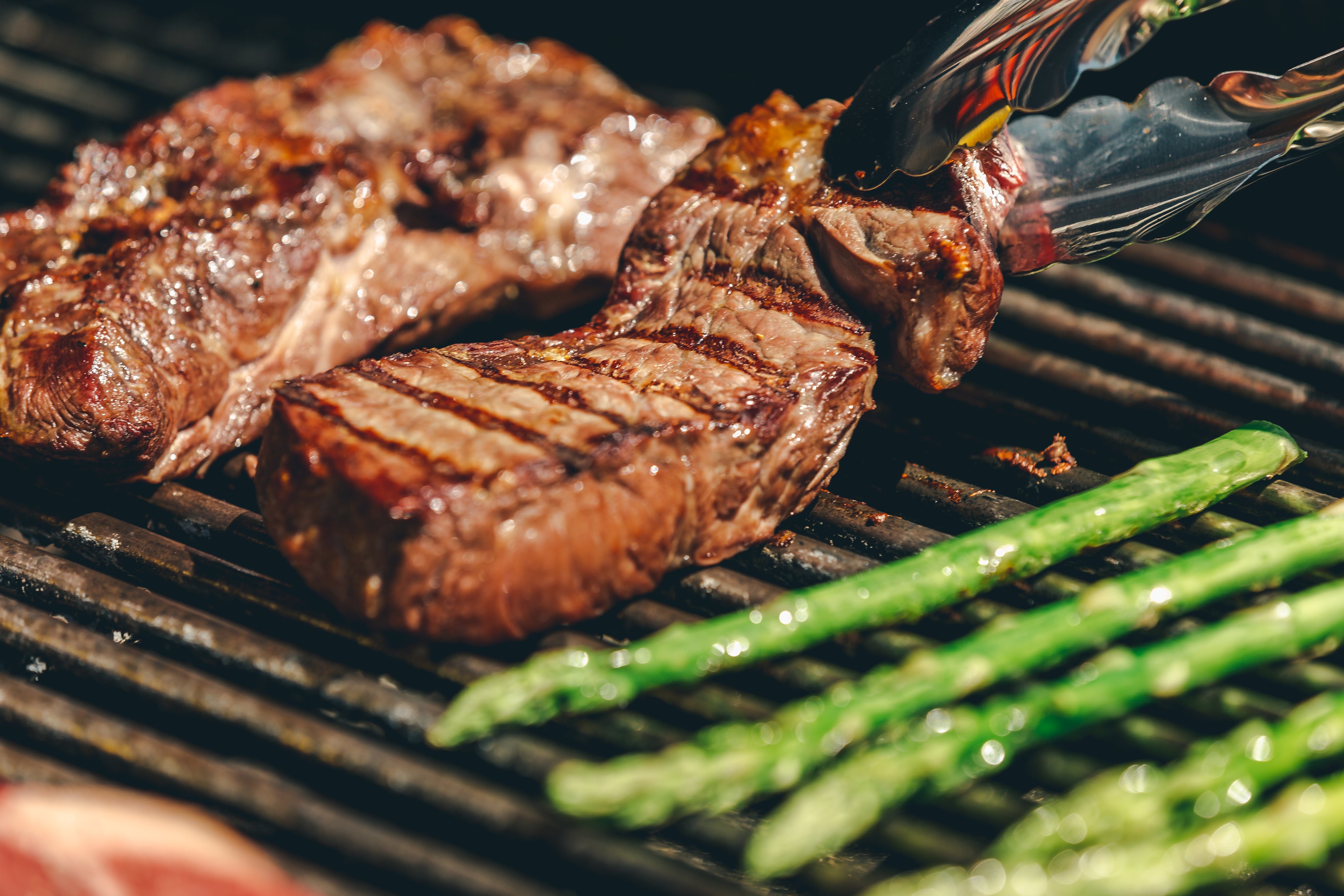 Grilled steak being turned with tongs on a barbecue grill, with asparagus cooking alongside