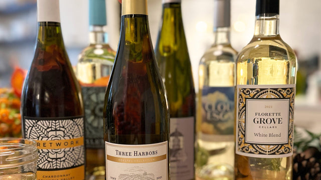 A selection of six Chardonnay wine bottles displayed on a table, with a hand reaching for one of the bottles. The bottles are labeled differently, indicating various brands.