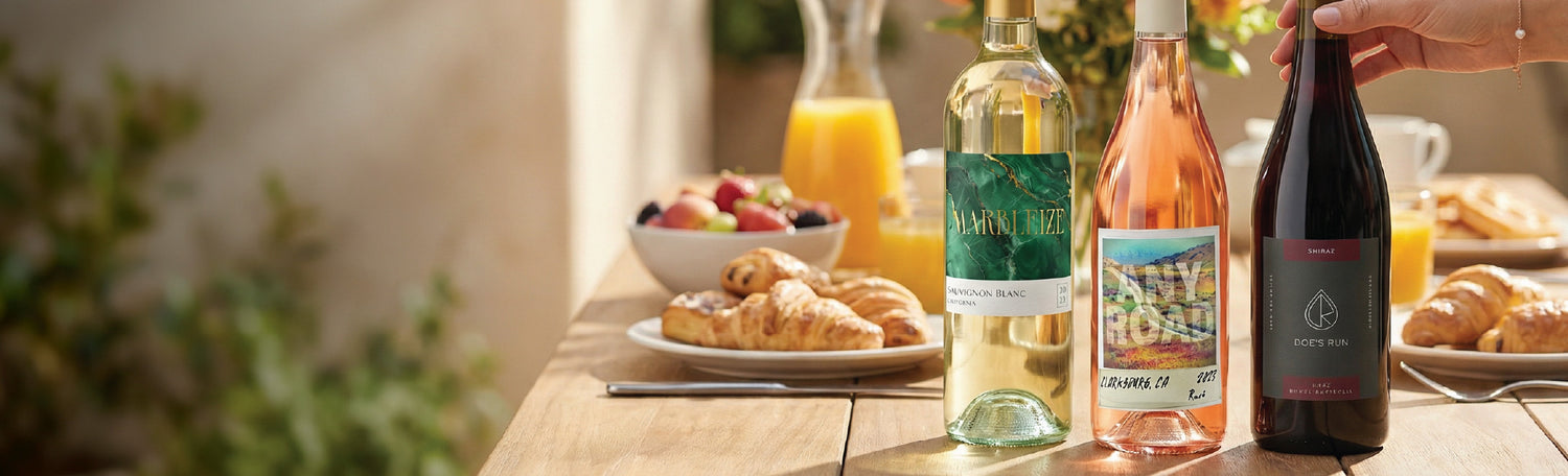 White, rosé, and red wine bottles on a brunch table with croissants, fresh fruit, and orange juice in the background.