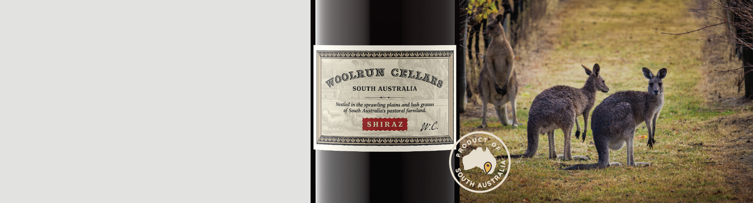 Wine bottle labeled ‘Woolrun Cellars Shiraz, South Australia’ next to three kangaroos standing in a grassy vineyard path.