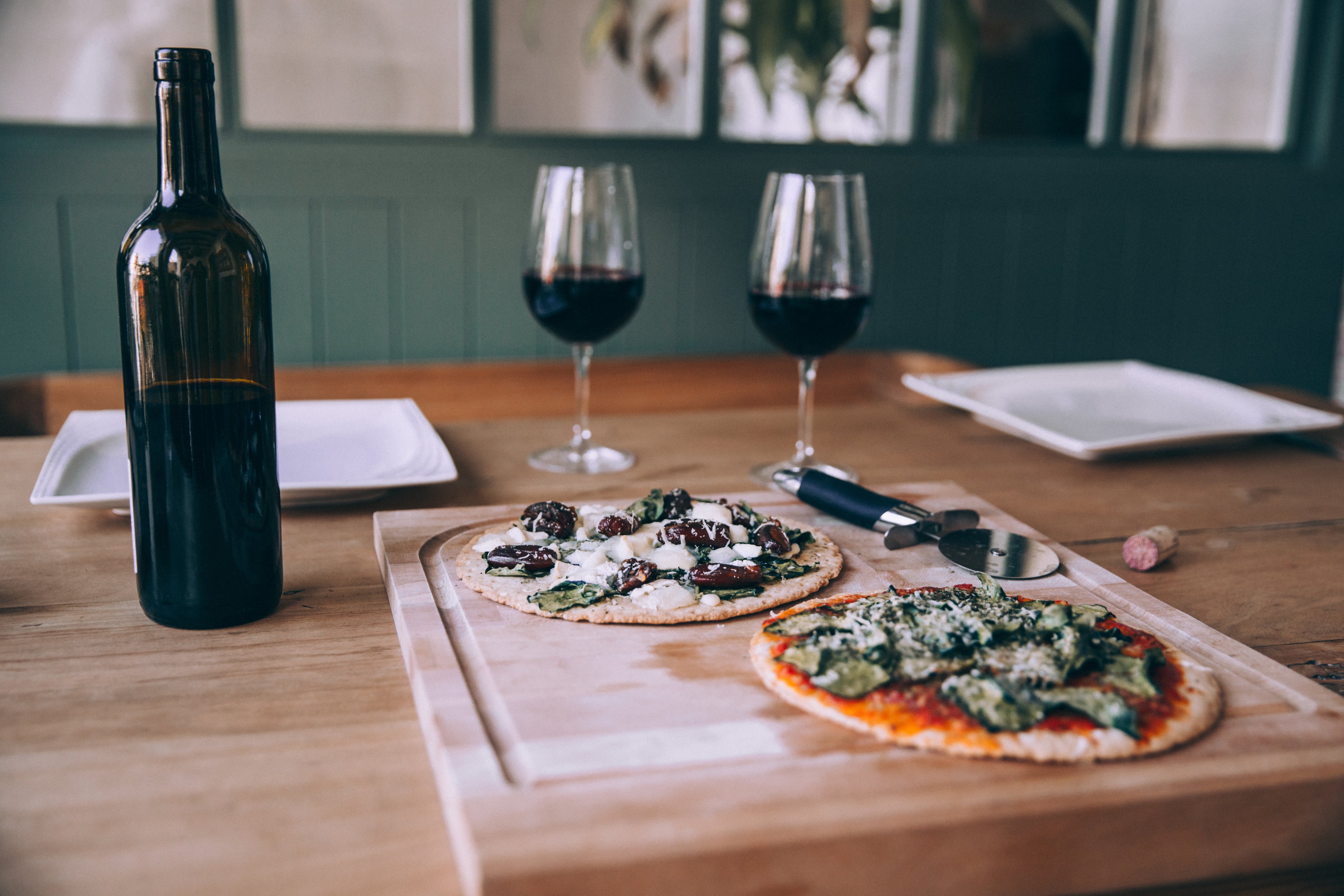 pizza on cutting board with red wine bottle and two glasses of red wine on table with white plates