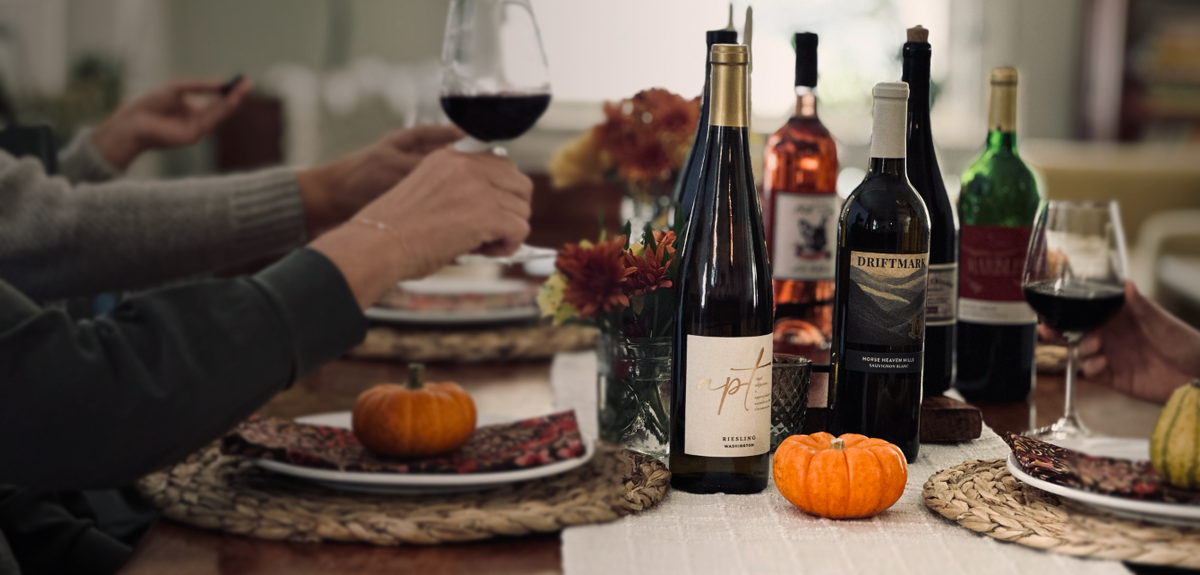 Wine bottles on a fall-themed dinner table with small pumpkins, flowers, and people holding glasses of red wine.